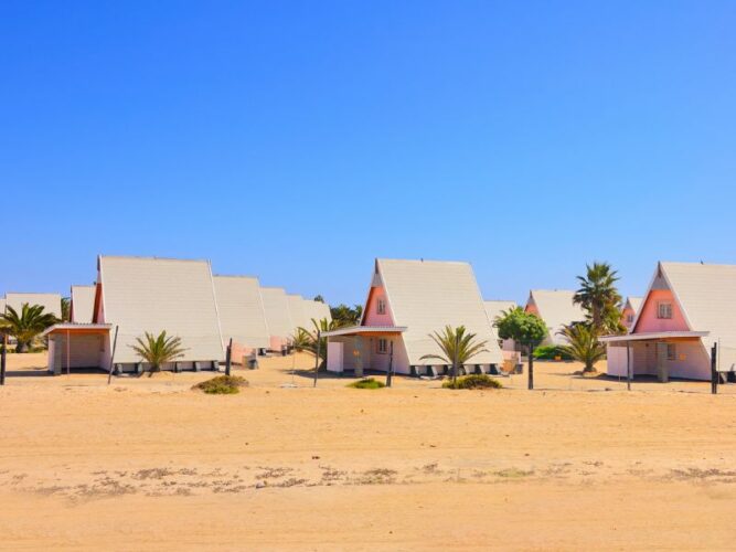 Quaint, triangular-roofed cottages line the sandy landscape in Swakopmund, Namibia, with palm trees punctuating the blue sky, capturing the charm of this coastal desert oasis.