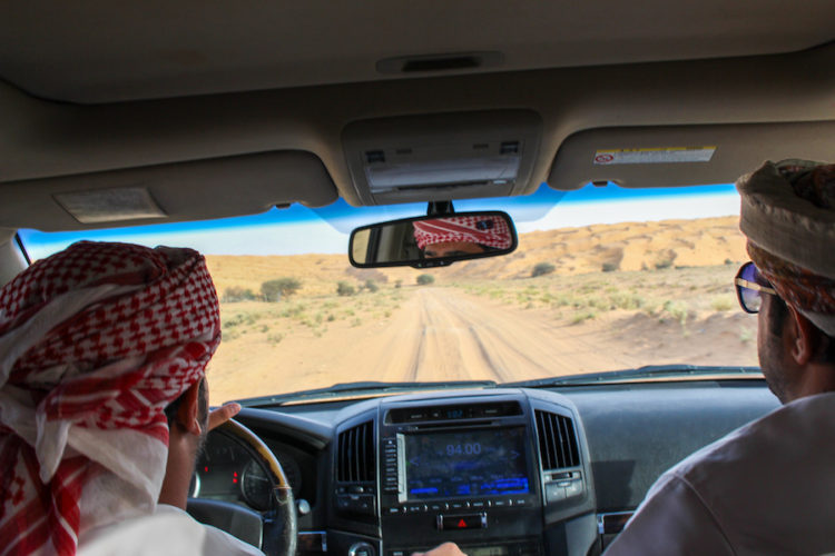 View from the backseat of an SUV with two men dressed in traditional Omani dress sat in the front driving through the desert on a sandy track on the edge of the wahiba sands