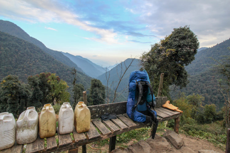 Blue Osprey backpack sitting on a wooden bench next to some plastic water containers with a view of mountains and valleys beyond