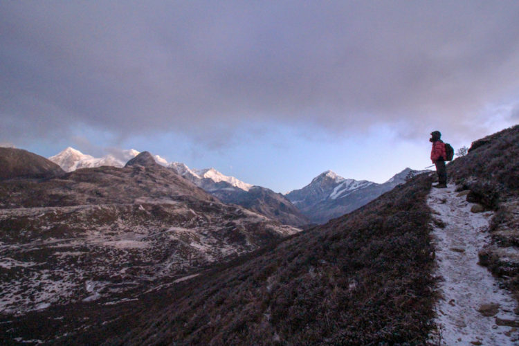 Man wearing a thickly padded jacket and hat on a frozen trekking trail in Sikkim looking across at the towering mountains