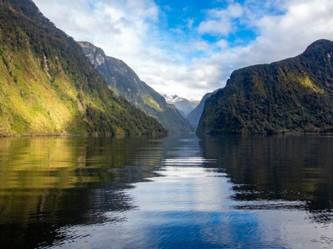 Calm waters of Doubtful Sound reflecting the lush green cliffs and distant snowy peaks under a partly cloudy sky in New Zealand