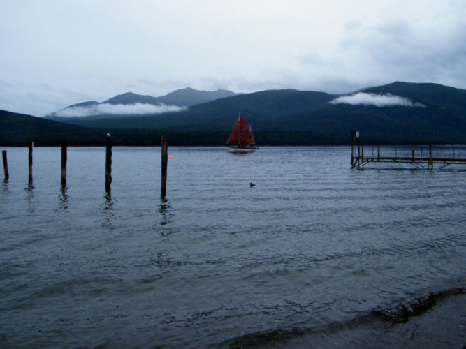 A moody photos of Lake Te Anau on a grey day with low level clouds and a boat with red sails in the middle of the lake