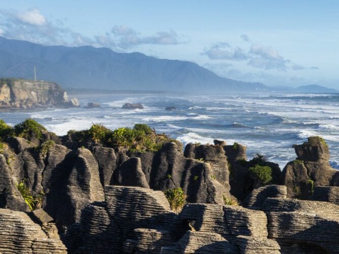 Punakaiki's famous Pancake Rocks and blowholes on New Zealand's wild West Coast, with layered limestone formations standing against the powerful Tasman Sea waves and a mountainous backdrop.