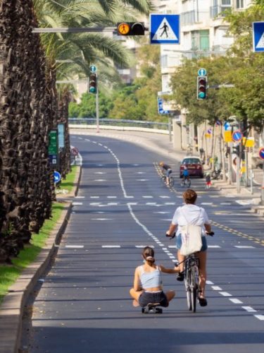 Woman sitting on a skateboard being pulled along the road by a cyclist in Tel Aviv
