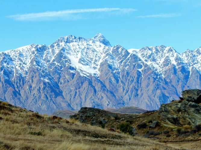 The Remarkables mountain range rising with jagged peaks covered in snow, overlooking a grassy landscape in the foreground