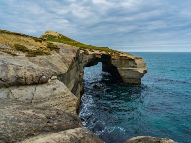 Dramatic natural archway carved into the rock at Tunnel Beach, near Dunedin, with turbulent ocean waters swirling below and a cloudy sky above