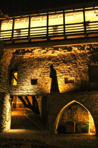 Creepy silhouette of a monk sculpture outside Kiek in de Kök at night with an old medieval archway illuminated by warm yellow lamps