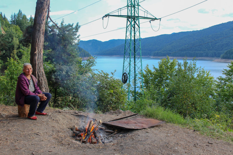 Woman sitting next to a fire with a scenic lake beyond in rural Romania