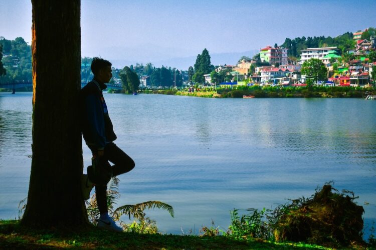 Man leaning against a tree next to the lake at Mirik with colourful buildings on the far shore