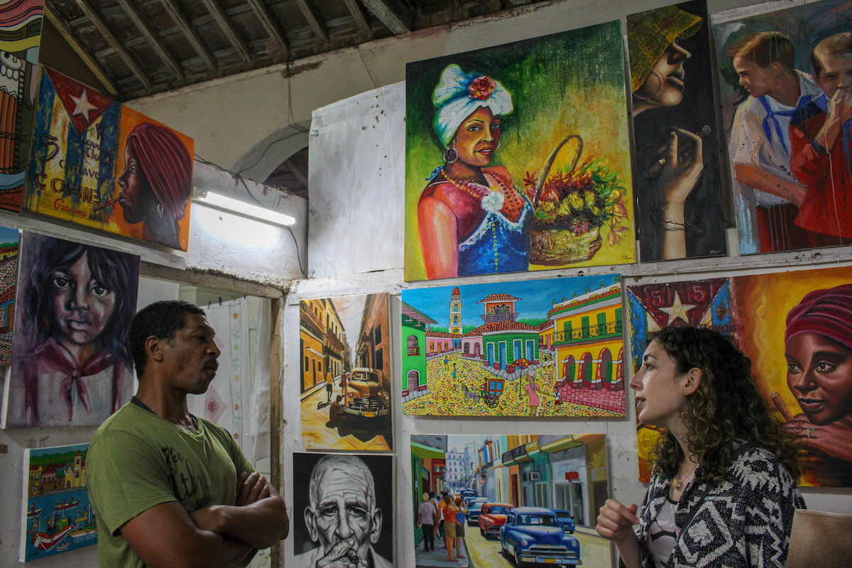 friendly cuban artist talking to a woman inside his studio with oil paintings on the walls