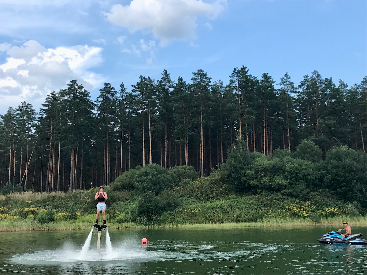 man flyboarding using a water powered hover board and a jet ski on a scenic lake in latvia with pine forests behind