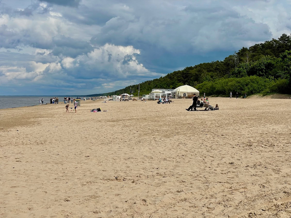 The wide, sandy stretch of Jūrmala Beach hosts small clusters of sunbathers, kids playing and a few white beach tents, with dense pine forest backing the dunes. Dramatic cumulus clouds roll above the calm, grey-blue Baltic Sea.