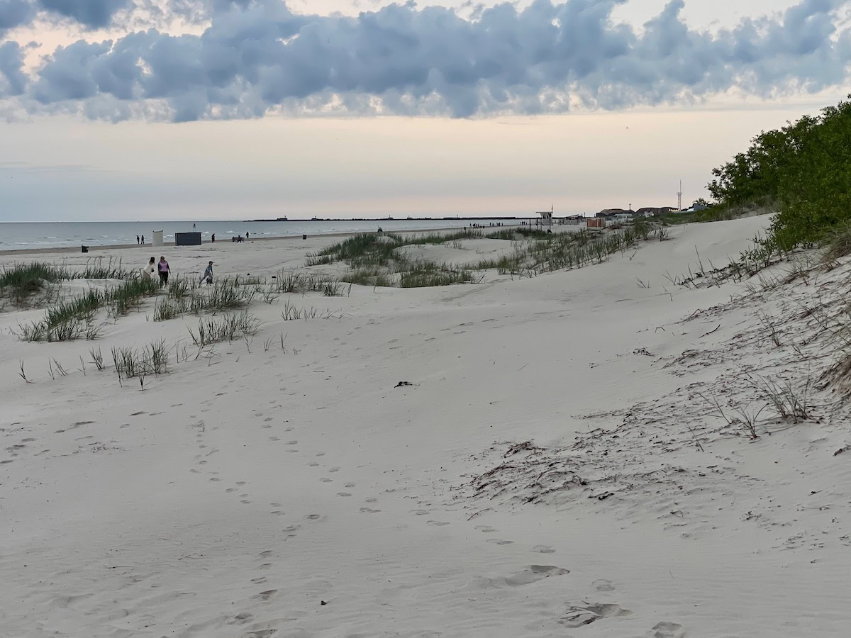 Soft grey-white sand dunes at Liepāja Beach slope gently to the Baltic Sea, dotted with wispy dune grasses and scattered footprints. A few evening strollers and children play near the shoreline beneath a pastel sky streaked with low, rolling clouds.
