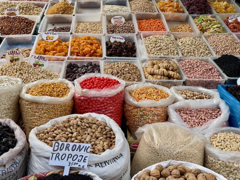 A colorful display of dried fruits, nuts, and legumes at Pazari i Ri market in Tirana, Albania. Large sacks and plastic containers are filled with almonds, cashews, dried apricots, figs, and beans, with handwritten labels indicating prices.