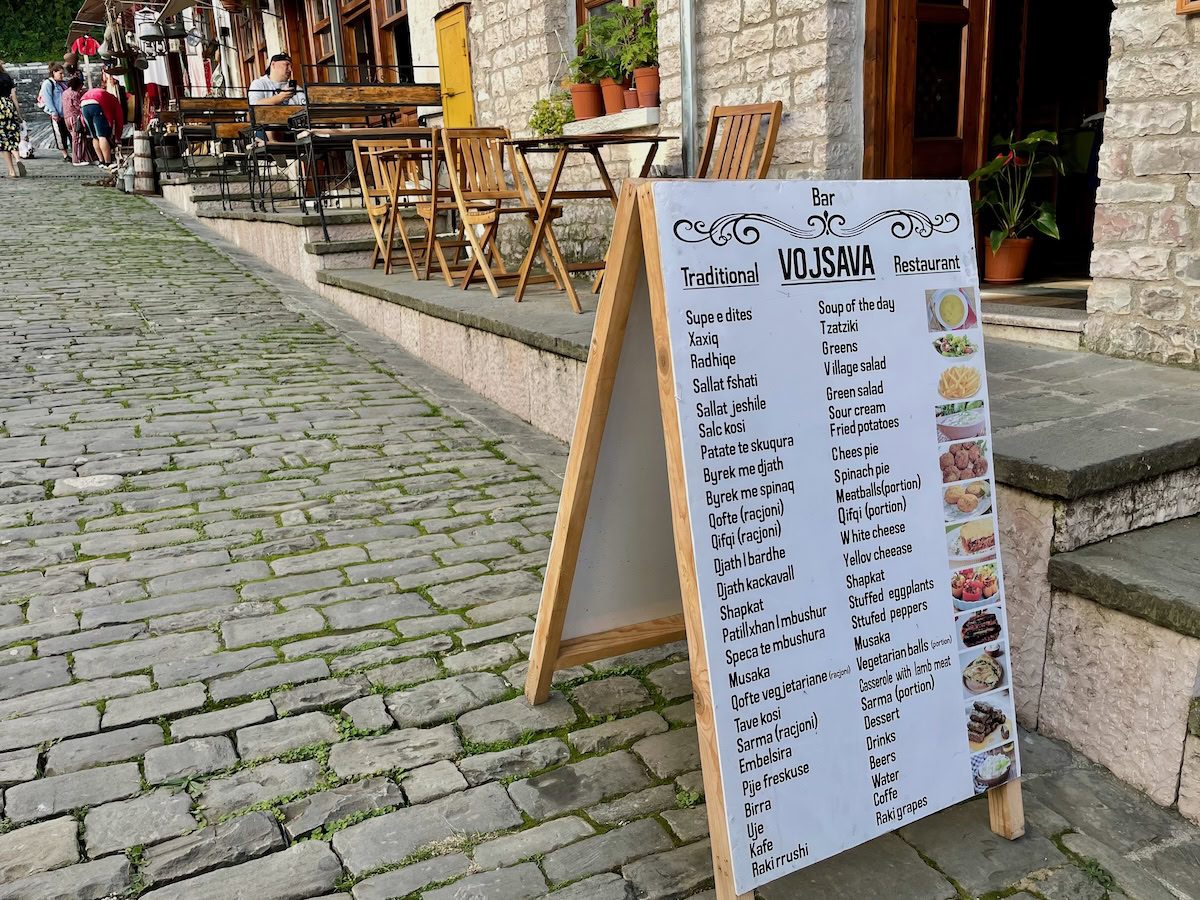 Street view of the Old Bazaar in Gjirokaster featuring cobblestone paths, outdoor cafés, and a menu board for Bar Vojsava listing traditional Albanian dishes. A lively spot and one of the top things to do in Gjirokaster for food and culture lovers.