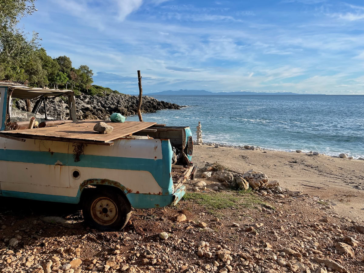 Old rusted van parked by a small beach with turquoise sea and rocky shoreline in southern Albania.