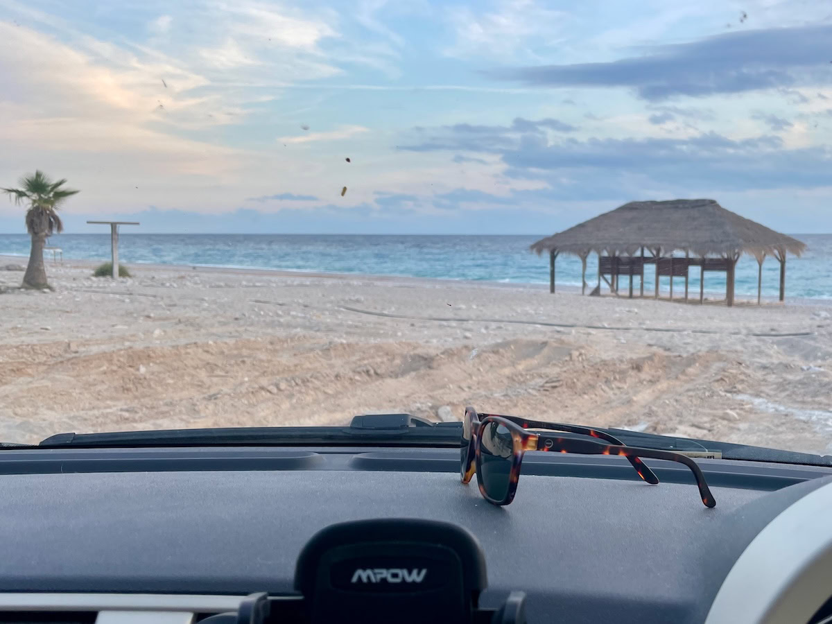 View from inside a parked car looking out to a sandy beach with a palm tree, wooden hut, and the sea in Albania.